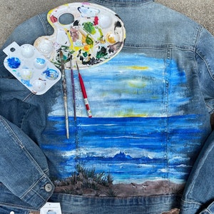 May include: A denim jacket with a painted seascape on the back. The painting features a blue sky, a blue ocean with waves, and a sandy beach with birds. There is a paint palette and paintbrushes on the left side of the jacket.