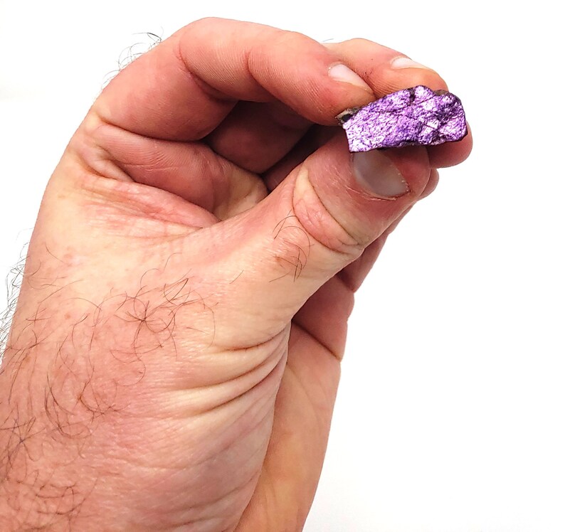 Pode incluir: Um close-up de uma amostra mineral de cor p&uacute;rpura e prateada. O mineral tem uma superf&iacute;cie &aacute;spera e irregular e um brilho met&aacute;lico.