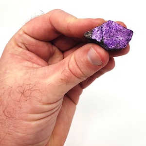 Pode incluir: Uma pequena rocha &aacute;spera, de cor roxa e cinza, com um brilho met&aacute;lico. A rocha est&aacute; sendo segurada em uma m&atilde;o.