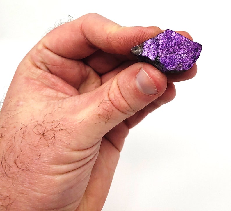 Pode incluir: Uma pequena rocha &aacute;spera, de cor roxa e cinza, com um brilho met&aacute;lico. A rocha est&aacute; sendo segurada em uma m&atilde;o.