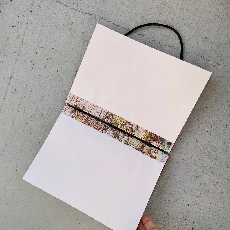 May include: A white notebook with a black elastic band closure. The notebook has a vintage world map design on the cover.