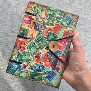 May include: A vintage-style journal cover with a collage of colorful postage stamps from Newfoundland. The stamps feature various denominations and designs, including a portrait of a man, a ship, and a lighthouse. The journal cover is bound with a black cord and has a brown leather button.