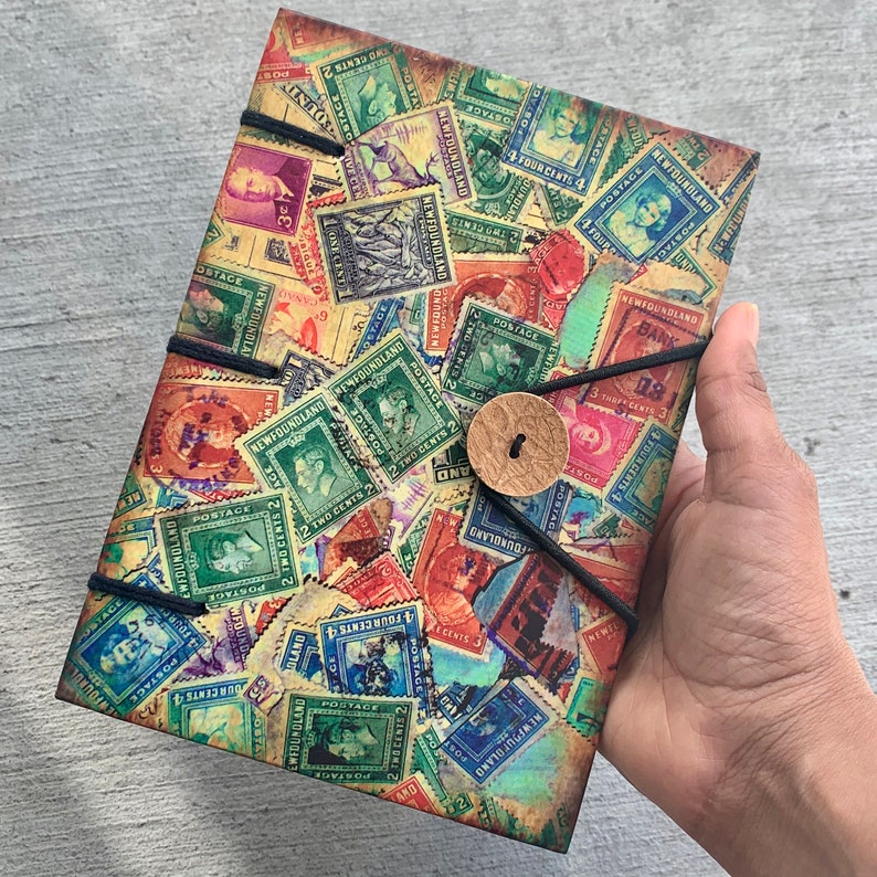 May include: A vintage-style journal cover with a collage of colorful postage stamps from Newfoundland. The stamps feature various denominations and designs, including a portrait of a man, a ship, and a lighthouse. The journal cover is bound with a black cord and has a brown leather button.