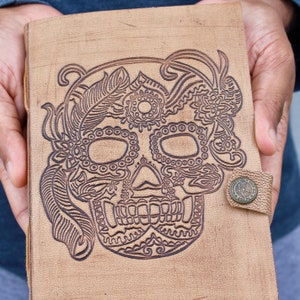May include: A tan leather journal featuring a carved sugar skull design on the cover, with floral and feather details. The journal is secured by a small metal clasp. The journal's dimensions are approximately 20 cm.