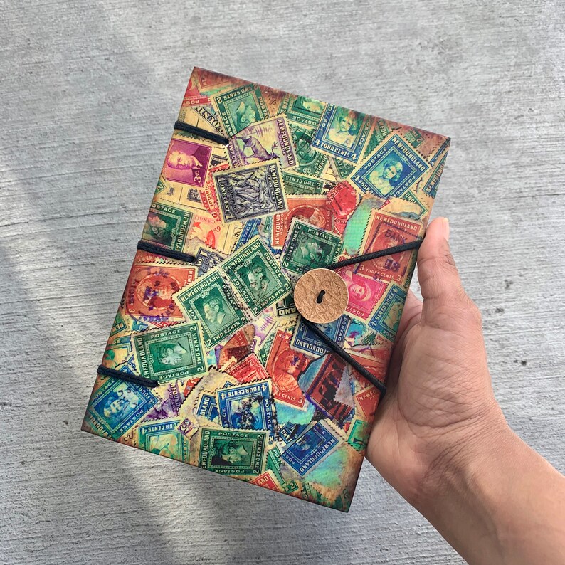 May include: A vintage-style journal with a cover made of old postage stamps from various countries. The cover is a collage of colorful stamps with images of people, animals, and landscapes. The journal has a brown leather strap with a button closure.