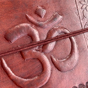 K&ouml;nnte beinhalten: Ein braunes Lederjournal mit einem gepr&auml;gten Om-Symbol und einem braunen Schnallverschluss.