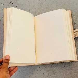 May include: An open blank journal with a tan leather cover and a brown leather strap. The journal is lying open on a gray surface.
