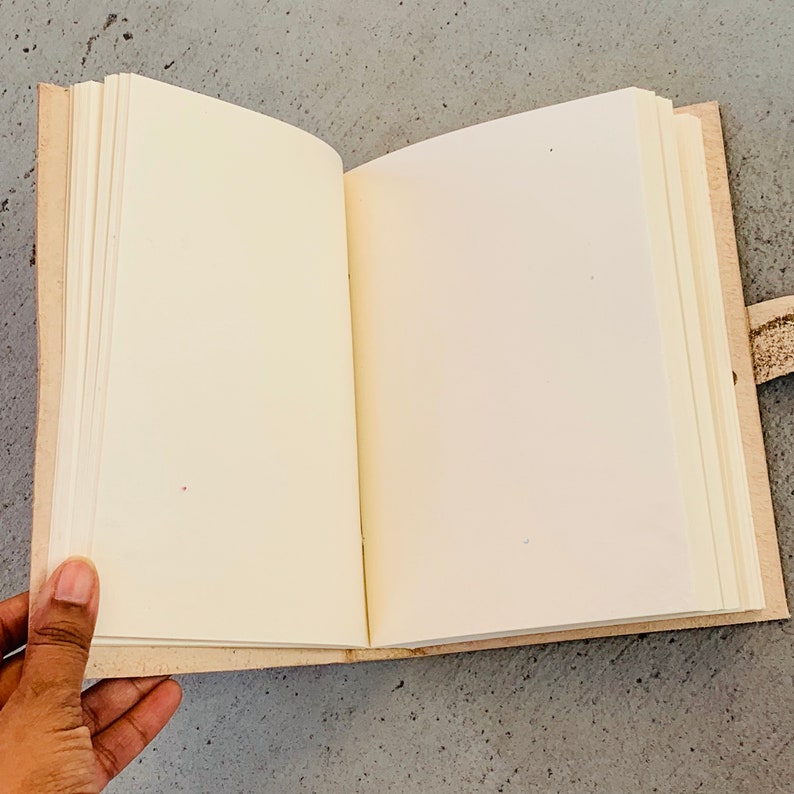 May include: An open blank journal with a tan leather cover and a brown leather strap. The journal is lying open on a gray surface.