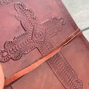 May include: A brown leather journal with a cross embossed on the cover. The cross is decorated with a geometric pattern and the journal has a brown leather strap.