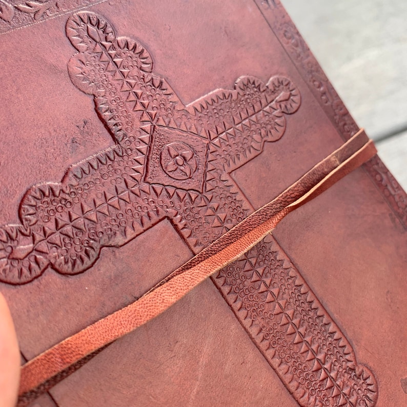 May include: A brown leather journal with a cross embossed on the cover. The cross is decorated with a geometric pattern and the journal has a brown leather strap.