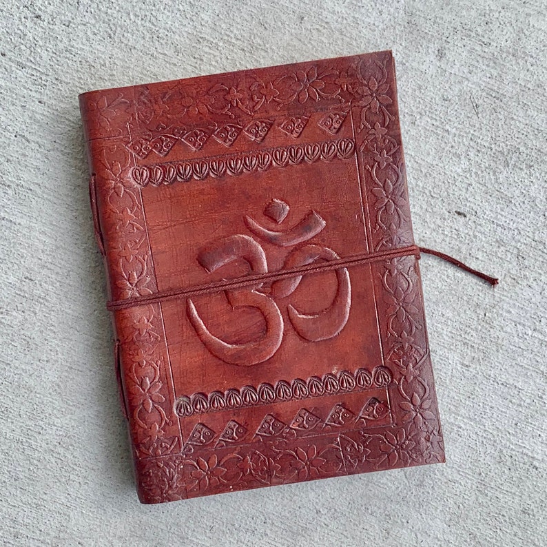 K&ouml;nnte beinhalten: Ein braunes Lederjournal mit einem gepr&auml;gten Om-Symbol und filigranen Blumenmustern. Das Journal hat einen Lederverschluss.