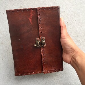 May include: A brown leather-bound journal with a brass clasp and decorative stitching. The journal is closed and held in a hand.