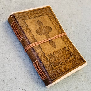 May include: A brown leather journal with a Celtic knot design embossed on the cover. The journal is closed and tied with a brown string.