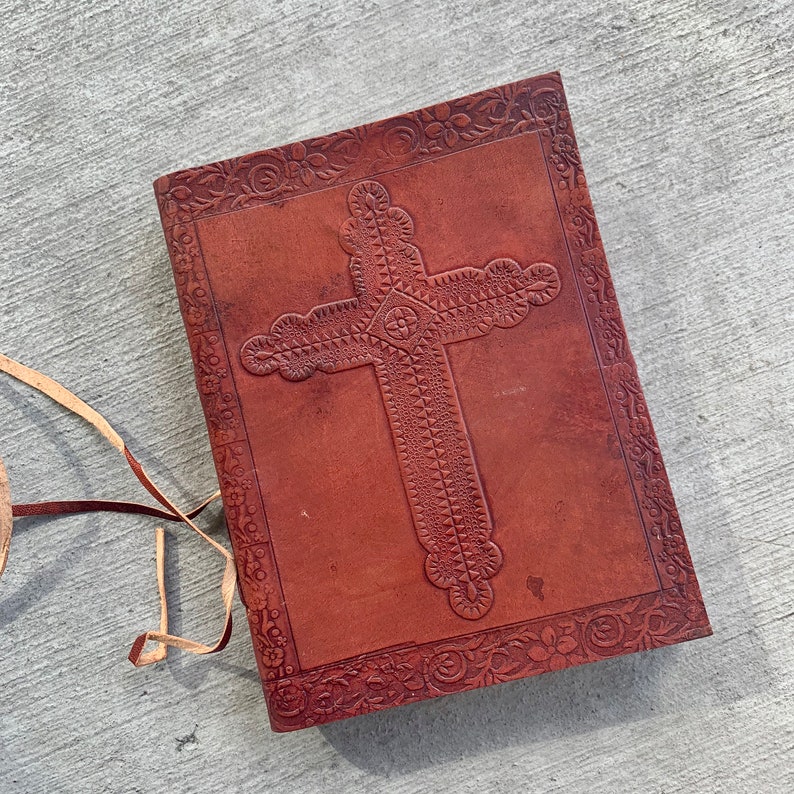 May include: A brown leather journal with a cross embossed on the front cover. The cross is surrounded by a floral design. The journal has a leather strap closure.