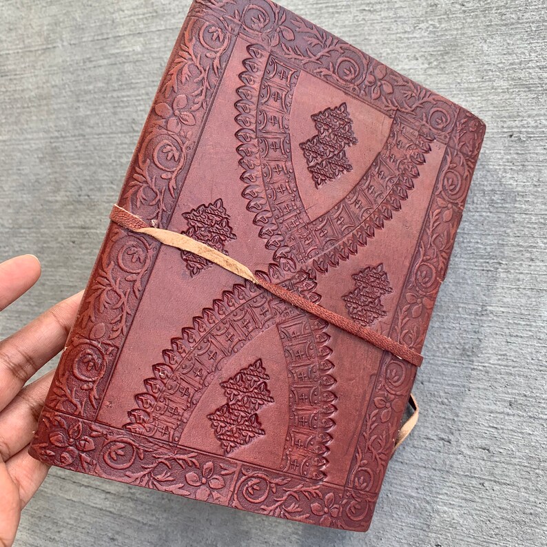 May include: A brown leather journal with intricate embossed floral and geometric patterns. The journal has a leather strap closure and is perfect for writing, journaling, or sketching.