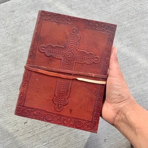 May include: A brown leather journal with a cross design embossed on the front cover. The journal has a leather strap closure and is decorated with floral patterns.