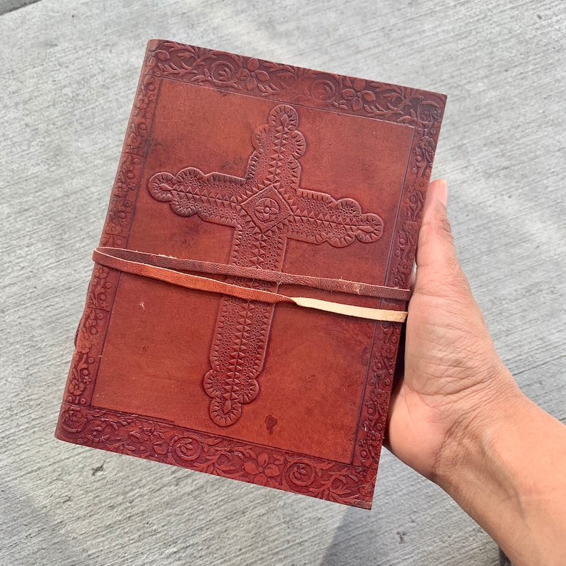 May include: A brown leather journal with a cross design embossed on the front cover. The journal has a leather strap closure and is decorated with floral patterns.