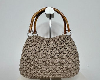 May include: A beige crocheted handbag with brown bamboo handles. The bag is displayed on a silver stand.