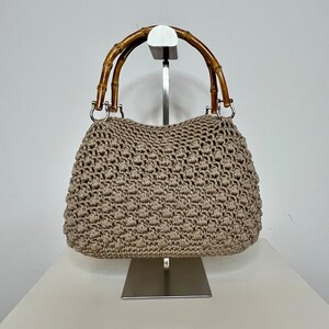 May include: A beige crocheted handbag with bamboo handles. The bag is displayed on a silver stand.