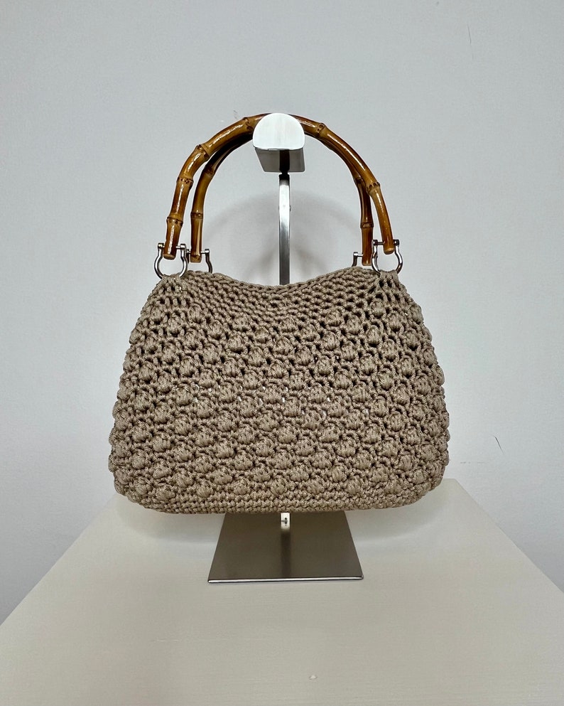 May include: A beige crocheted handbag with bamboo handles. The bag is displayed on a silver stand.
