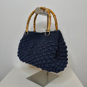 May include: A navy blue crocheted handbag with bamboo handles. The bag is displayed on a silver stand.