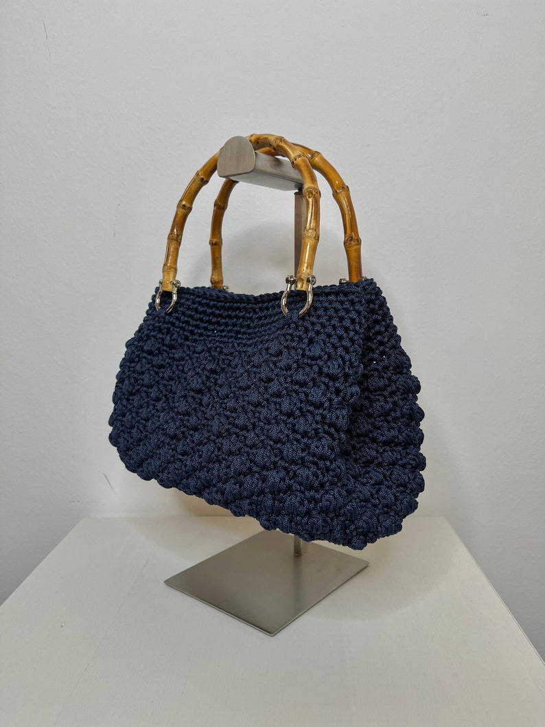 May include: A navy blue crocheted handbag with bamboo handles. The bag is displayed on a silver stand.