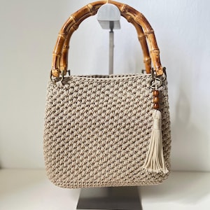 May include: A beige crocheted handbag with a bamboo handle and a tassel. The bag is displayed on a silver stand.