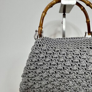 May include: A grey crocheted handbag with a bamboo handle. The bag is on a display stand.