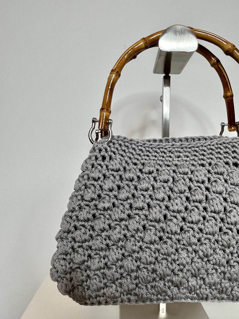 May include: A grey crocheted handbag with a bamboo handle. The bag is on a display stand.