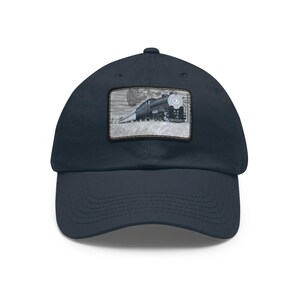 May include: A navy blue baseball cap with a rectangular patch showing a black and white illustration of a steam train. The train is on a track with a cloudy sky. The cap has a curved peak.