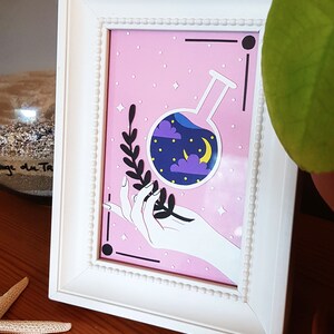 Peut inclure: Un cadre blanc avec un fond rose représentant une main tenant un flacon en verre avec un ciel nocturne à l'intérieur. Le flacon a une lune croissante et des nuages. Le fond est recouvert de points blancs.