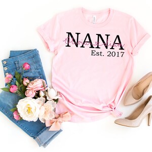 以下が含まれることがあります： 白地にピンクのTシャツに、黒い文字で「NANA Olivia & Liam Est. 2017」と書いてあります。Tシャツは、青いジーンズとピンクと白の花束と一緒に、白い背景の上に平らに置かれています。