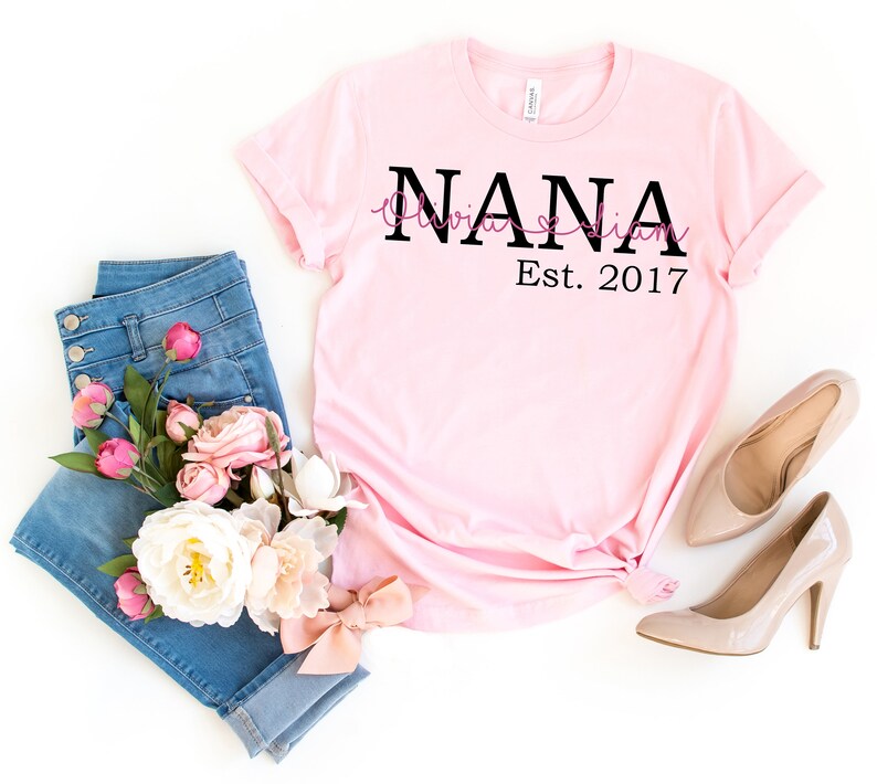 以下が含まれることがあります： 白地にピンクのTシャツに、黒い文字で「NANA Olivia & Liam Est. 2017」と書いてあります。Tシャツは、青いジーンズとピンクと白の花束と一緒に、白い背景の上に平らに置かれています。