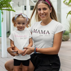 以下が含まれることがあります： 「MAMA Est. 2017」と書かれた白いTシャツ。白いTシャツに「Olivia Est. 2017」と書かれた子供は、女性の腕を握っている。