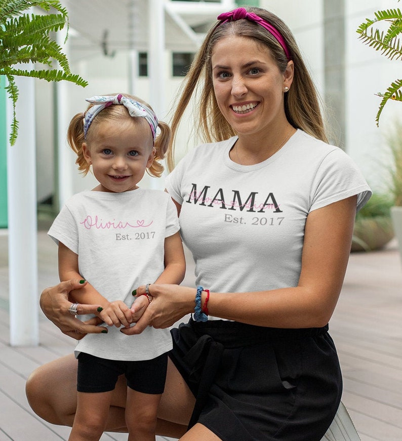以下が含まれることがあります： 「MAMA Est. 2017」と書かれた白いTシャツ。白いTシャツに「Olivia Est. 2017」と書かれた子供は、女性の腕を握っている。