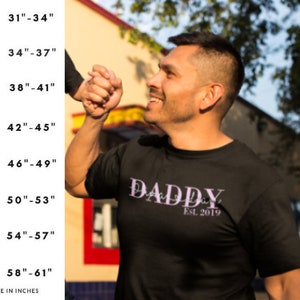 以下が含まれることがあります： 紫色の書体で「DADDY」と書かれた黒いTシャツで、「Est. 2019」という文字が下にあります。インチで胸囲のサイズ表が示されています。