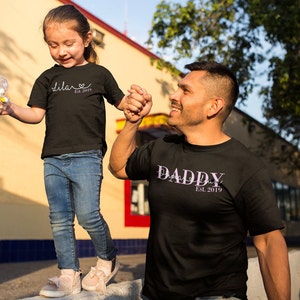 以下が含まれることがあります： 父親と娘がお揃いの黒いTシャツを着ています。娘のTシャツには「Lila Est. 2019」と書いてあり、父親のTシャツには「Daddy Est. 2019」と書いてあります。