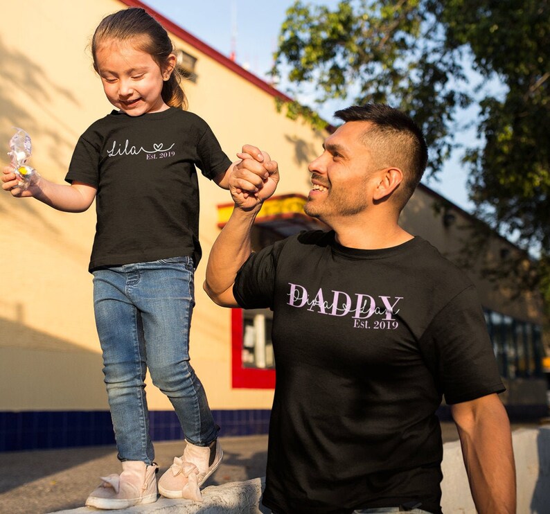 以下が含まれることがあります： 父親と娘がお揃いの黒いTシャツを着ています。娘のTシャツには「Lila Est. 2019」と書いてあり、父親のTシャツには「Daddy Est. 2019」と書いてあります。