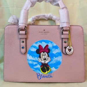 Op de afbeelding: Een roze Kate Spade handtas met een handgeschilderd ontwerp van Minnie Mouse in de wolken. De tas heeft een bovenste handvat en een gepersonaliseerde naam, "Brenda", geschreven in blauw onder het ontwerp. Een kleine hanger met de woorden "Handgeschilderd door Billie Jean" hangt aan het handvat.