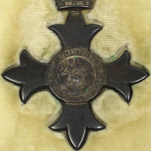 Può includere: Una medaglia scura a forma di croce con un nastro viola, presentata in una custodia foderata di velluto. La medaglia presenta un emblema circolare centrale e un dettaglio a corona nella parte superiore, che suggerisce un oggetto storico o commemorativo.