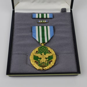 Puede incluir: Una medalla de oro con una cinta verde y azul en una caja de terciopelo negro. La medalla presenta un águila con las alas extendidas y un escudo con estrellas.