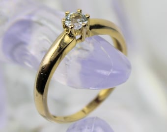 Op de afbeelding: Een gouden ring met een enkele, heldere edelsteen in een kraalbezetting.