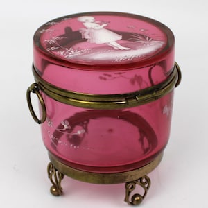 Könnte beinhalten: Eine Vintage-Schmuckdose aus rubinrotem Glas mit Messingrahmen und verzierten Füßen. Der Deckel zeigt eine gemalte Szene einer Figur. Die Dose ist zylindrisch, mit einem Klappdeckel und einem Griff. Die Dose ist etwa 10 cm hoch.
