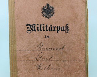 Puede incluir: Un antiguo Militärpaß alemán, o pasaporte militar, con una cubierta rosa descolorida. La cubierta presenta el texto "Militärpaß" y otro texto alemán, junto con un emblema. El documento es de 1904.
