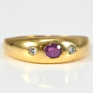 Könnte beinhalten: Ein goldener Ring mit einem zentralen Rubin und zwei kleineren Diamanten auf beiden Seiten. Der Ring hat ein glattes, abgerundetes Band. Die Edelsteine sind in einem klassischen Stil gefasst. Schmuck.