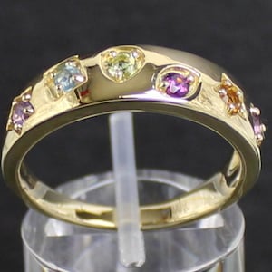 Könnte beinhalten: Ein goldener Ring mit einer Reihe farbiger Edelsteine. Der Ring hat ein leicht gewölbtes Band, und die Edelsteine sind oben eingesetzt. Die Edelsteine sind in verschiedenen Farben gehalten, darunter Lila, Orange, Grün und Blau.