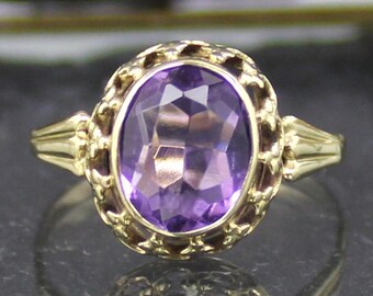Puede incluir: Un anillo de oro con una gran piedra preciosa ovalada de color púrpura. La piedra preciosa está rodeada por un borde decorativo de piedras más pequeñas y oscuras. La banda del anillo tiene un diseño clásico con detalles sutiles.