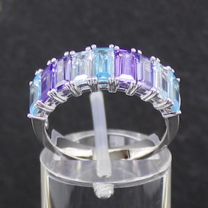 Könnte beinhalten: Ein silberner Ring mit einer Reihe rechteckiger Edelsteine in Blau- und Lilatönen. Der Ring wird auf einem transparenten Ständer präsentiert, der das Design und die Farbvariationen hervorhebt. Die Edelsteine sind in einem Band angeordnet.