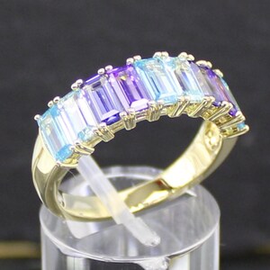 Könnte beinhalten: Ein goldener Ring mit einer Reihe rechteckiger Edelsteine in Blau- und Lilatönen. Der Ring wird auf einem transparenten Ständer vor dunklem Hintergrund präsentiert. Die Edelsteine sind in einem Farbverlauf angeordnet.