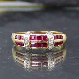 Op de afbeelding: Een gouden ring met een kanaalzetting van vierkant geslepen rode robijnen. Het midden van de ring wordt geaccentueerd door twee verticale rijen kleine, heldere, rond geslepen stenen, wat een opvallend brugontwerp vormt over de rode band.